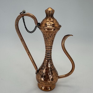May include: A copper-colored, hammered metal pitcher with a long, curved spout and handle. The pitcher has a bulbous base, a slender neck, and a lid attached by a chain. The surface has a textured, dimpled pattern, and the overall design is ornate.