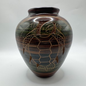 May include: A decorative ceramic vase with a turtle design. The vase is brown with green and blue stripes and a carved turtle motif. The vase has a wide body and a narrow neck.
