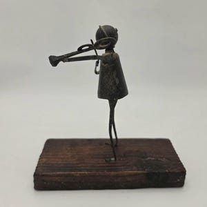 May include: A small, metal sculpture of a person playing a violin. The figure is dark gray and stands on a rectangular, wooden base. The violin is held up to the figure's face, and the bow is in the right hand. The sculpture has a rustic, handmade appearance.