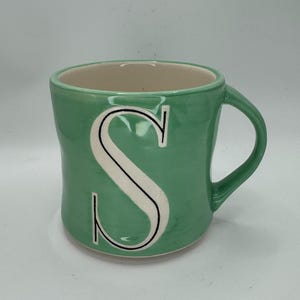 May include: A green ceramic mug with a large white letter "S" outlined in black. The mug has a curved handle and a light beige interior. The exterior has a textured appearance.