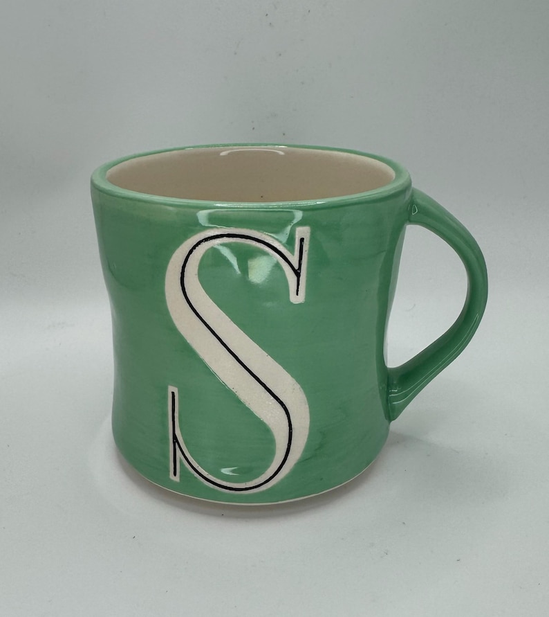 May include: A green ceramic mug with a large white letter "S" outlined in black. The mug has a curved handle and a light beige interior. The exterior has a textured appearance.