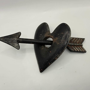 May include: A black metal heart pierced by an arrow. The heart has a hole in the center and a rustic finish. The arrow has a pointed tip and fletching, with a dark finish. The piece is likely a decorative item.