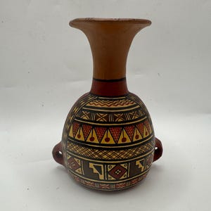 May include: A terracotta vase with a flared rim and two small side handles. The bulbous body features intricate geometric patterns in black, red, and yellow, including zigzags, triangles, and cross-hatching, painted across the surface.