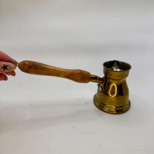 May include: A brass coffee pot with a wooden handle. The pot has a rounded base and a narrow spout. The handle is a light brown color. The pot is designed for making Turkish coffee.