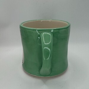 May include: A green ceramic mug with a curved handle. The mug has a light green exterior and a beige interior. The mug is sitting on a white surface.