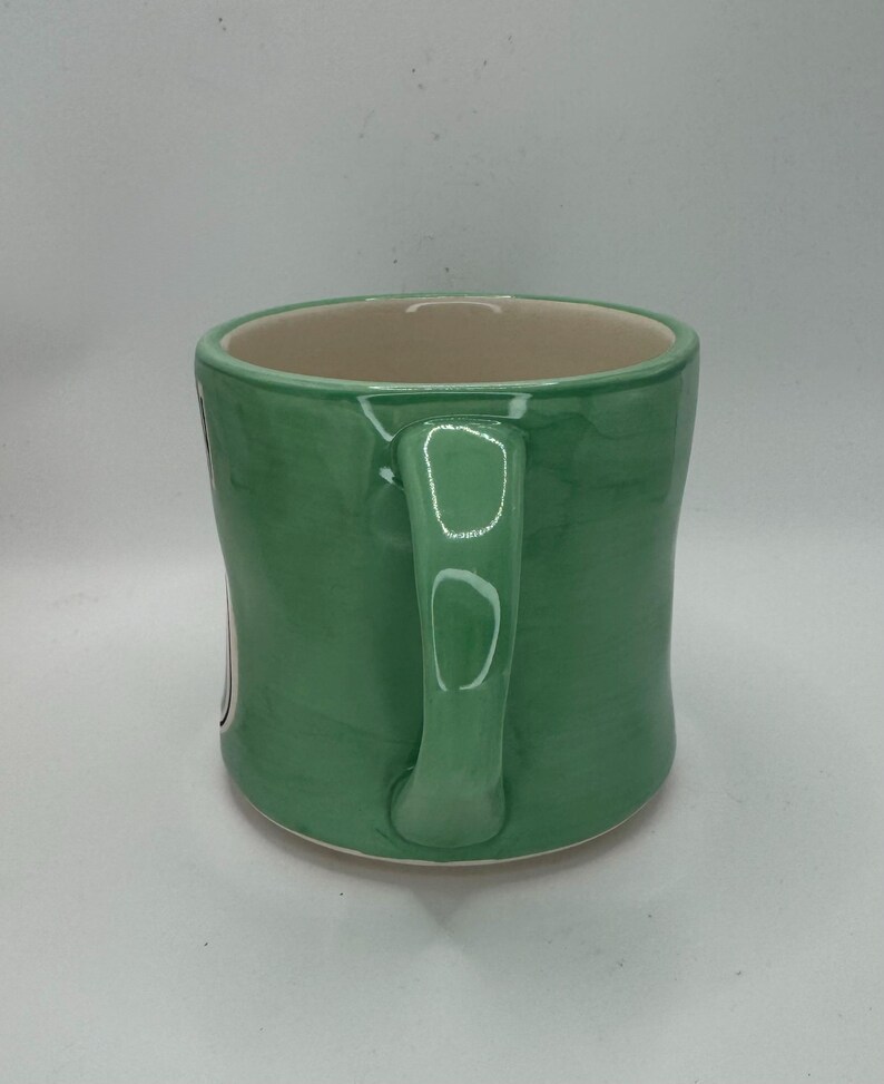 May include: A green ceramic mug with a curved handle. The mug has a light green exterior and a beige interior. The mug is sitting on a white surface.