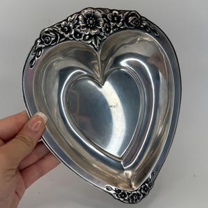 May include: A heart-shaped, silver-colored dish with a floral border. The dish has a smooth, reflective surface and a detailed floral design around the rim. The dish is held by a person's hand.
