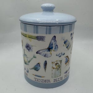 May include: A cylindrical ceramic canister with a matching lid, featuring a light blue and white striped rim. The body displays watercolor illustrations of a blue butterfly, birds, a watering can, and lavender, with the text TENDER BLUE FLOWERS.