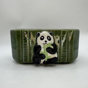 May include: A green ceramic planter with a panda bear design. The panda is black and white, holding a green bamboo shoot. The planter has a rectangular shape with bamboo-like detailing.