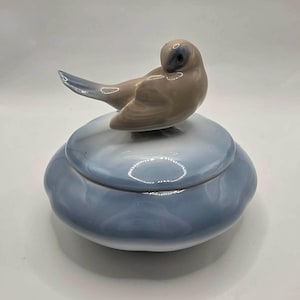 May include: A round ceramic trinket box with a glossy finish, featuring a light blue gradient base and a matching lid. A small, sculpted ceramic bird in shades of beige, brown, and soft blue is perched on top of the lid, facing toward the left side.