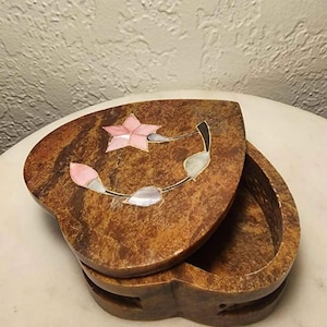 May include: Heart-shaped jewelry box with a hinged lid. The box is brown with a floral design inlaid with pink and white mother-of-pearl. The box is open, revealing the interior. The box is sitting on a white surface.
