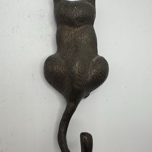 May include: Bronze-colored cat-shaped wall hook. The hook is designed in the shape of a cat sitting with its back to the viewer, with a curved tail forming the hook. The cat has pointed ears and a textured surface.