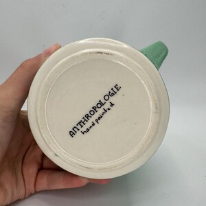 May include: A white ceramic mug with a green handle. The bottom of the mug features the text "ANTHROPOLOGIE hand painted" in black lettering. The mug is held by a person.