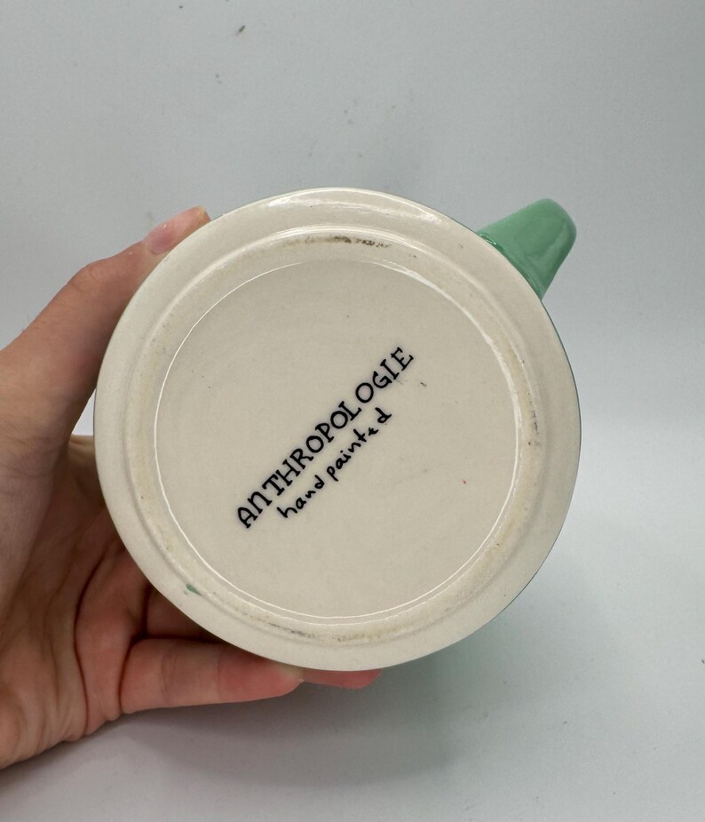 May include: A white ceramic mug with a green handle. The bottom of the mug features the text "ANTHROPOLOGIE hand painted" in black lettering. The mug is held by a person.