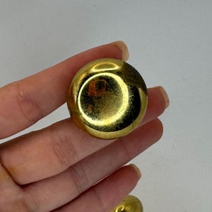 May include: A close-up of a gold-colored, round earring. The earring has a shiny, reflective surface. The earring is held in a hand.