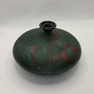 May include: A low-profile, disc-shaped ceramic vase with a dark, textured forest green finish. The surface features subtle, mottled reddish-brown undertones and a smooth, circular opening at the top with a distinct, slightly raised black rim.