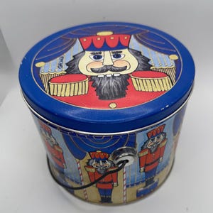 May include: A blue metal tin with a lid featuring a nutcracker design. The tin has a handle and is decorated with illustrations of nutcrackers in red coats and blue curtains. The lid has a large nutcracker illustration.