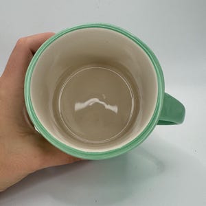 May include: A ceramic mug with a light green handle and rim. The interior is a light beige color. The mug is held by a hand.