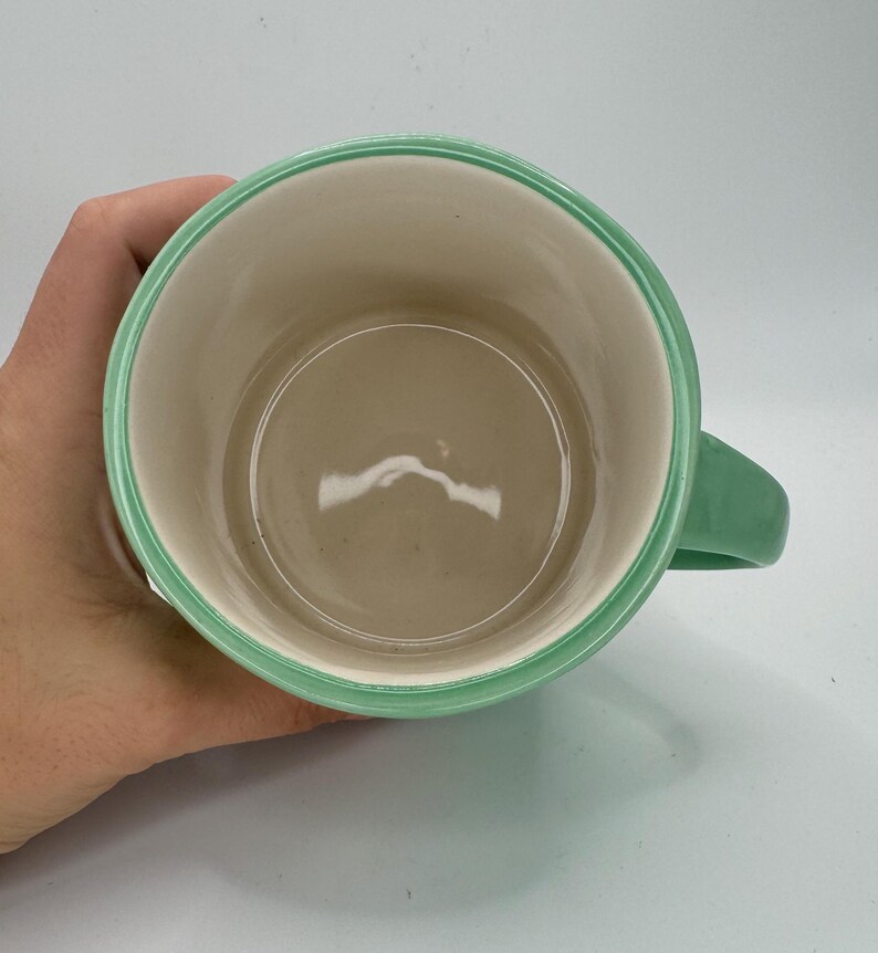 May include: A ceramic mug with a light green handle and rim. The interior is a light beige color. The mug is held by a hand.