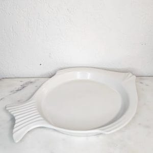 May include: A white ceramic serving dish in the shape of a fish. The dish has a round center and a tail fin with vertical lines. The dish is on a marble surface.