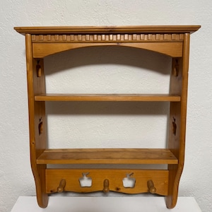 May include: A light brown wooden wall shelf with three shelves and three pegs. The top of the shelf has a decorative carved design. The sides have apple-shaped cutouts. The shelf is made of wood and has a natural finish.