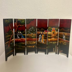 May include: A six-panel folding screen with an Asian-inspired design. Each panel features a dark red background with painted scenes of figures, trees, and buildings. The screen is made of wood and has a black frame.