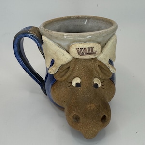 May include: A ceramic mug with a moose design. The mug is primarily brown with a blue handle and accents. The word "VAIL" is printed on the front. The mug has a unique, handcrafted appearance.