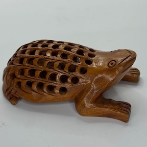 May include: A detailed wooden frog sculpture with a carved, openwork design on its back. The frog is a warm brown color and has detailed features, including eyes and legs. The sculpture is a decorative item.