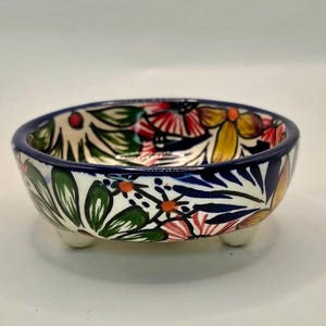 May include: A small, round ceramic bowl with a floral design. The bowl features a white base with colorful flowers in red, yellow, and pink, and green leaves. It has a dark blue rim and sits on three small, white feet.
