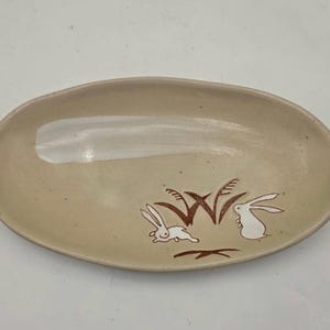 May include: Oval-shaped ceramic dish in a beige tone. The dish features a brown illustration of two rabbits and tall grass. The design is simple and elegant, suitable for serving small portions or as a decorative piece.