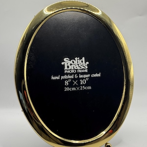 May include: Oval, solid brass photo frame. The frame is hand polished and lacquer coated. The frame is 8" x 10" (20cm x 25cm) and has a black interior. The text "Solid Brass PHOTO FRAME" is visible.