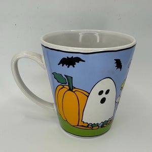 May include: A white ceramic Halloween mug with a light blue background. The mug features a cartoon pumpkin, ghost, and bats. The mug has a white handle and a white interior.