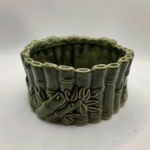 May include: A round, olive green ceramic planter featuring a molded, raised relief design of bamboo stalks and leaves encircling the exterior. The vessel has a glossy, glazed finish and an open top, showcasing a hollow interior for holding items.