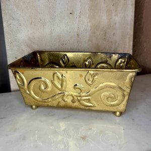 May include: A rectangular brass planter with ornate floral scrollwork and small feet. The planter has a golden hue and is likely made of metal. The design features stylized leaves and swirls, adding a decorative element to the piece.