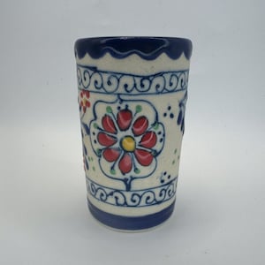May include: A cylindrical ceramic tumbler featuring a cream-colored base with hand-painted floral motifs in red, yellow, and green. The rim and base are accented with a solid dark blue band, and the body displays intricate blue scrollwork.