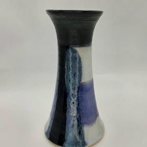 May include: A ceramic vase with a flared rim and a tapered body. The surface features a black glaze at the top, transitioning into vertical bands of white, deep blue, and a textured, mottled light blue glaze that runs down the center of the piece.