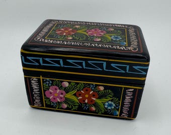 May include: A black rectangular wooden box with hand-painted floral designs in pink, red, and blue. The box has a geometric pattern in blue and yellow. The box is likely used for storing small items.