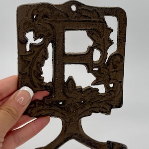 May include: A decorative, cast iron wall hook in a dark brown finish. The hook is shaped like the letter "F" with ornate floral detailing and a double hook at the bottom. The hook measures approximately 6 inches in height.
