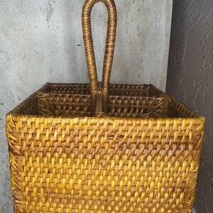 May include: A square, woven rattan basket with a curved handle. The basket is a warm, golden-brown color and has a textured surface. The handle is made of the same material and is attached to the top of the basket.