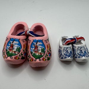 May include: Two pairs of Dutch wooden clogs. One pair is pink with a windmill scene, the other is white with a blue windmill design. Both pairs have a red, white, and blue ribbon detail.