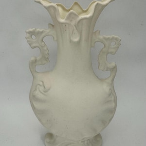 May include: Off-white ceramic vase with a flared top and ornate handles. The vase features a textured surface and a decorative base with leaf-like details. The handles have a dragon-like design.