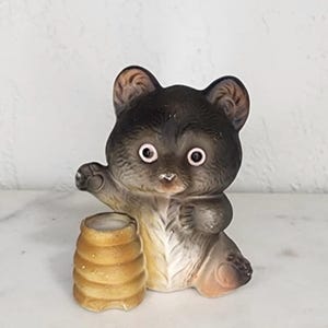 May include: A vintage ceramic bear figurine with a honey pot. The bear is primarily dark brown with lighter brown accents on the ears and paws. It has large, round eyes and is holding a small, tan honey pot. The figurine is on a white surface.