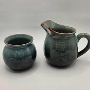 May include: A ceramic creamer pitcher and a matching small bowl featuring a glossy glaze in shades of deep blue, teal, and charcoal gray. The pitcher has a curved handle and a pouring spout, while the bowl has a rounded, open-top design.
