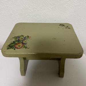 May include: A small, square, sage green wooden step stool with a floral design on the top. The flowers are in shades of red, purple, and yellow with green leaves. The stool has two legs on each side.