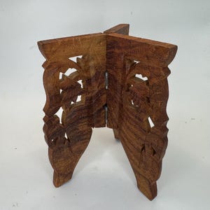 May include: A small, foldable wooden stand with intricate carved floral designs. The stand is made of dark brown wood and has three legs. The stand is designed to hold a small object. The stand is approximately 6 inches tall.