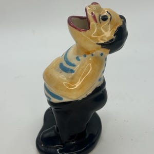 May include: A vintage ceramic figurine of a man with an open mouth, wearing black pants and a yellow shirt with blue stripes. The figurine has a black head and is standing on a black base.