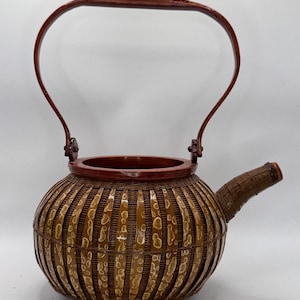 May include: A handcrafted, round bamboo teapot with a curved handle and spout. The teapot features a woven design with a reddish-brown rim and handle, and is supported by three small feet. The bamboo has a natural, light brown color with darker accents.