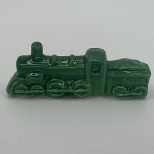 May include: A small, green ceramic train figurine. The train has detailed features including wheels, a smokestack, and a coal car. The word "WADE" is visible on the side of the coal car. The figurine is set against a plain white background.