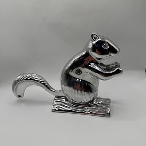 May include: A shiny, silver-colored metal nutcracker shaped like a squirrel. The squirrel is depicted in a seated position on a textured base that resembles a log, with its tail curved upward and its front paws held near its chest.