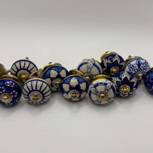 May include: A collection of ceramic cabinet knobs in various designs. The knobs feature a blue and white color scheme with floral and geometric patterns. Each knob has a brass-colored base.